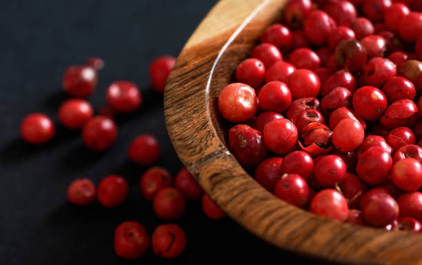 whole pink red peppercorns in small wooden bowl, some scattered on black gray paper, closeup view from above - pink pepper imagens e fotografias de stock
