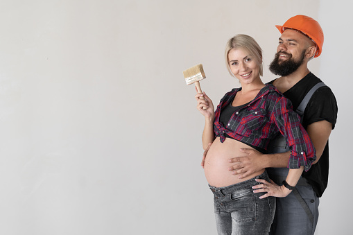 A man hugs his pregnant wife. work uniform and orange safety helmet. A woman holds a paintbrush in her hands. Place for text