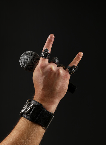 Close up man hand with metal rings and bracelet showing devil horns rock gesture sign, holding microphone over black background, side view
