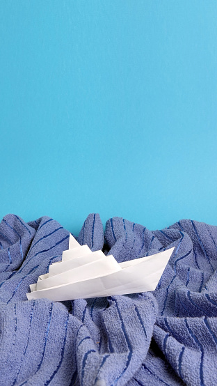 This is a white paper ship toy on blue towel waves abstract concept photo of turbulent sea or ocean.