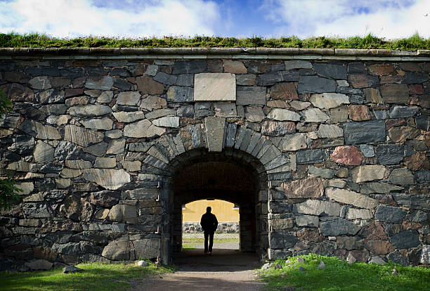 Suomenlinna Fortress. Closeup, Royal Gate gray stone ramparts, covered by weeds, under gray cloudscape. Green tree foliage
