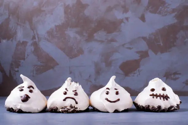 Funny meringue ghosts with funny faces on gray table. Halloween dessert. Copy space