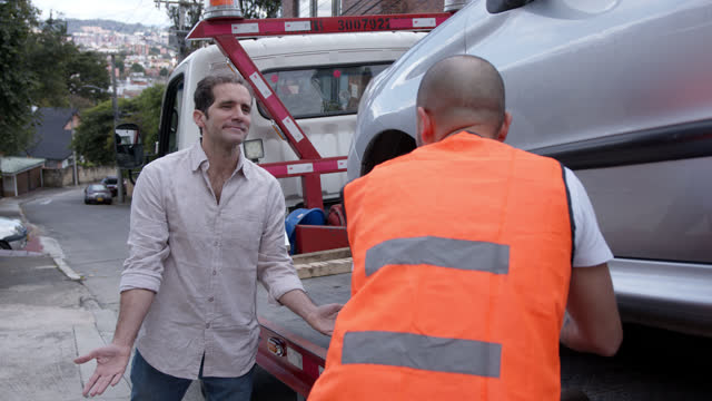 Latin American man discussing with a tow truck worker while he is securing car on platform