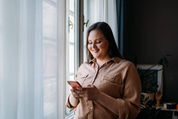 Beautiful Plus Size Woman Standing next to a Window while Browsing the Internet on her Smartphone