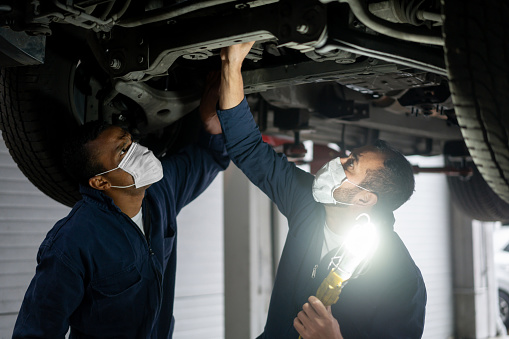 Team of Latin American mechanics wearing facemask while working at a garage fixing a car on a lift and using a lamp to look at the chassis during the COVID-19 pandemic
