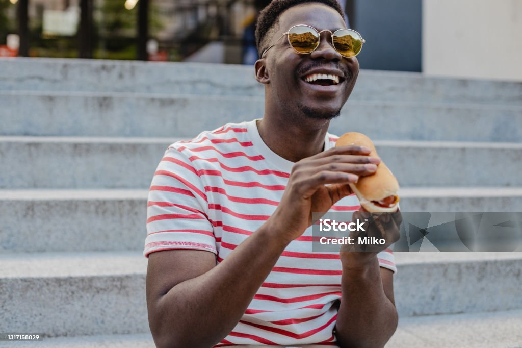 Young African-American man is eating hot dog and smiling A young modern Black man is eating a hot dog and smiling Eating Stock Photo