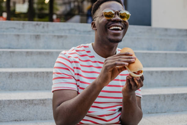 Young African-American man is eating hot dog and smiling A young modern Black man is eating a hot dog and smiling black people eating stock pictures, royalty-free photos & images