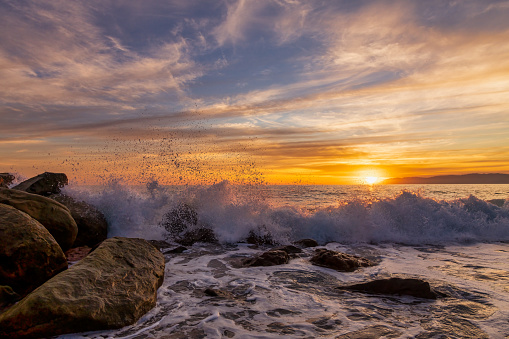A Wave Is Breaking And Splashing On Sea Rocks As The Sun Sets On the Ocean Horizon