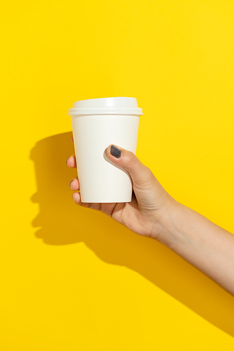 Hand holding disposable paper cup with plastic lid on yellow background