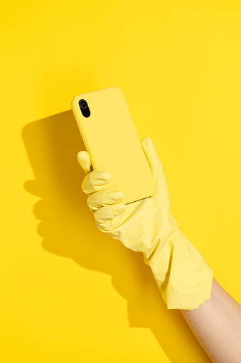 Hand wearing yellow rubber household cleaning gloves holding phone on yellow background