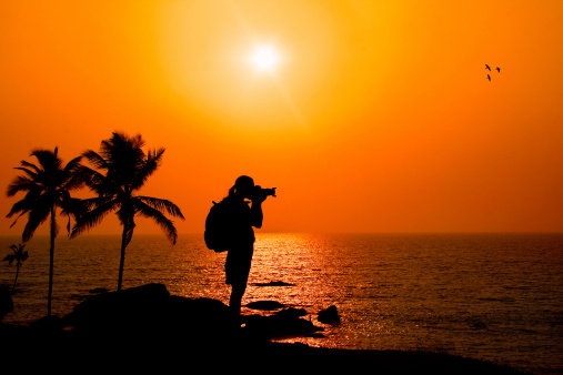 Photographer silhouette shooting sea outdoors on the rock cliff near palm trees at sunset background