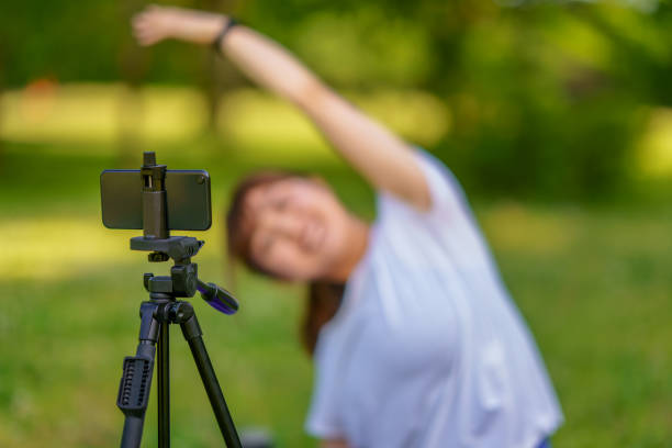 A female yoga instructor is using a smart phone on a tripod and teaching online class in nature.
