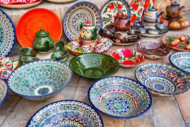 Typical colorful handcrafted traditional uzbek pottery plates and tea pot and tea cup ceramics on the floor at bazaar - market stall in the Medina of Bukhara. Handicraft Pottery in Bukhara - Buxoro - Бухорo, Silk Road, Khorezm Region, Uzbekistan, Central Asia.