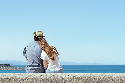 Couple sitting backwards while looking to the horizon at the shore in a sunny day.