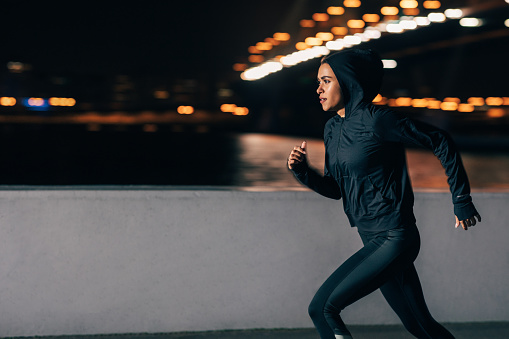 Side view of a woman in hoodie running at night. Middle east female jogger exercising outdoors.
