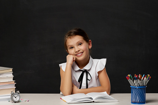 happy schoolgirl daydreaming and sitting at table in the classroom on chalkboard background. Back to school.