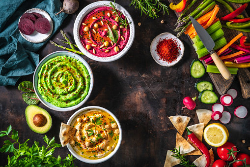 Hummus Three bowl of chickpeas, avocado and beetroot with cut vegetables for dip on dark wooden table background