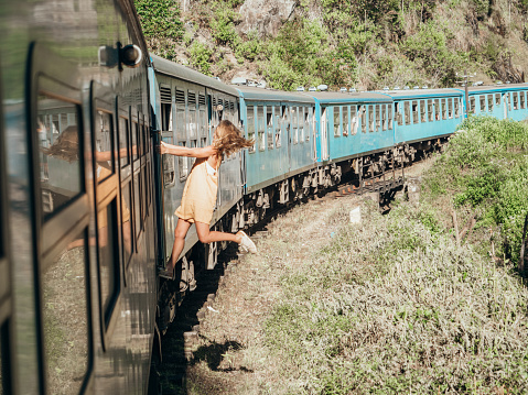 Traveling young woman hanging outside blue train door while riding on the famous Ella Kandy journey in Sri Lanka. People transportation famous places. Girl enjoying train ride in tea plantations