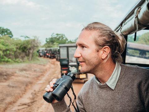 Happy young man on luxury safari searching for wild animals in the jungle using binoculars.Man in moving vehicle 4x4 looking for wildlife in national park