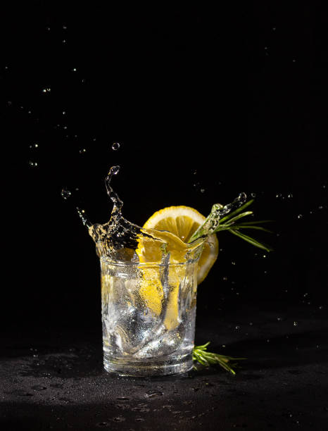 bevanda alcolica incolore in un bicchiere con una fetta di limone e rosmarino, splash. vodka su sfondo nero. - colors isolated white fruit foto e immagini stock