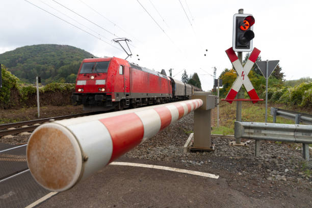 geschlossenes bahnübergangstor und rote warnleuchte mit einem güterzug im hintergrund - railroad crossing train railroad track road sign stock-fotos und bilder