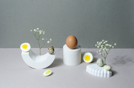 Modern still life concept with chicken, quail eggs and Gypsophila flowers on white stands and podiums over gray background. Minimalist style.