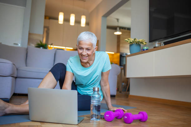 Senior woman sitting on the floor on exercise mat in front of laptop and training at home. Senior woman sitting on the floor on exercise mat in front of laptop and training at home. Beautiful senior woman doing stretching exercise while sitting on yoga mat at home. grey hair on floor stock pictures, royalty-free photos & images