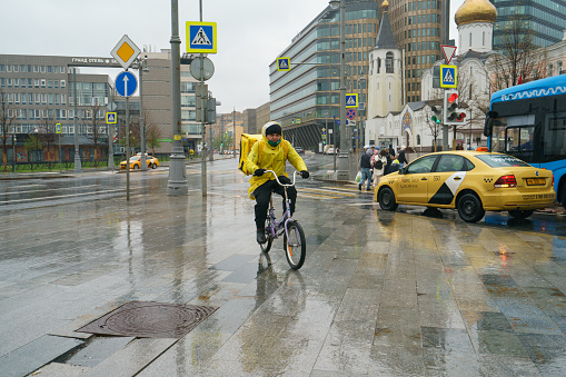 Moscow, Russia - May 8, 2021: Rainy day in Moscow. Delivery man cycling during coronavirus pandemic. He wear, use, put on protective face mask. Lifestyle and occupation concepts.