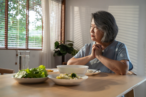 Asian Elderly retired grandmother stay at home with painful face sitting alone on eating table in house. Depressed mature Senior old woman upset feeling unhappy, lonely and missing her family at home.