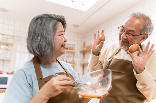 Asian Senior elderly couple standing in kitchen at house feeling happy and enjoy retirement life together. Loving Older grandparent cooking food with smiling face. Relationship and activity at home.