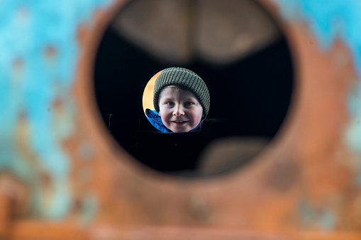 A young boy at a bottle bank is looking through a hole on one side of the bottle bank. This is shot through the other side of the bottle bank and only his face is visible.