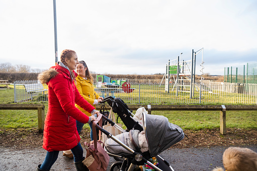 A side-view shot of two young caucasian mothers leaving the park with their pushchairs, they are wrapped up in warm clothing on a cold day and the young boy is running.
