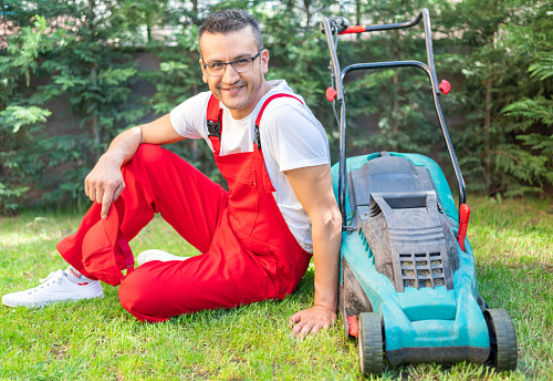 A portrait of gardener with a lawn mower is cutting grass in backyard.  A worker is cleaning in the garden.