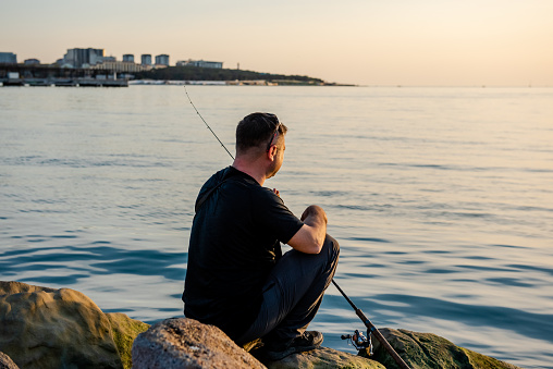A fisherman sits on a rock by the sea with a fishing rod at sunset. Single sea fishing. Day at the beach. Copy space for text