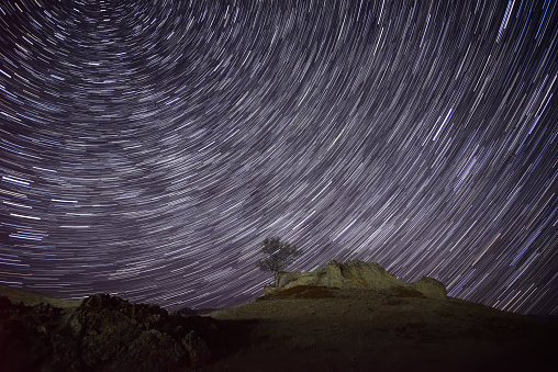 Night landscape with a tree and rocks on the top in the mountains against the background of a starry sky with traces of stars revolving around the polar star, in the form of circular tracks