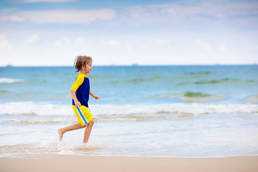 Kids playing on tropical beach. Children swim and play at sea on summer family vacation. Sand and water fun, sun protection. Little child running and jumping at ocean shore.