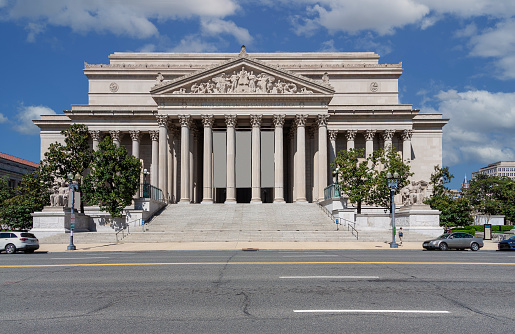 The National Archives Building, Washington DC, USA. Blue Sky with Puffy clouds, Street with Parked Cars, and Green Trees are in the image. Wide angle lens.\n.