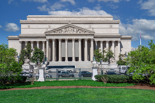 The National Archives Building, Washington DC, USA. Blue Sky with Puffy clouds, Green Lawn, Street with Parked Cars, and Trees are in the image.\n.