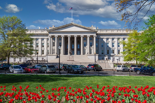 Treasury Department Building, Washington DC, USA. Red Flowers, Blue Sky with Puffy clouds and Green Lawn and Trees are in this image.\n.