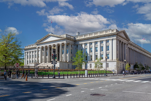 Treasury Department Building, Washington DC, USA. Blue Sky with Puffy clouds, Street, Sidewalk, Green Lawn and Trees are in the image.\n.