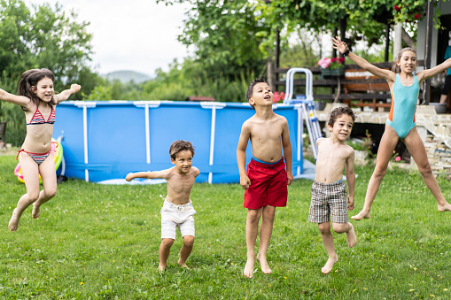 Children bouncing around outside on the grass, wearing swimming costumes, in a garden with a swimming pool behind