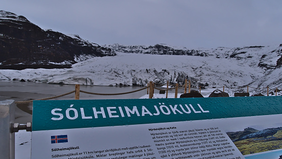 Sólheimajökull, Iceland - 03-24-2021: View of glacier Solheimajokull, part of Mýrdalsjökull, from viewpoint with information sign in front on cloudy day in winter season. Focus on sign.