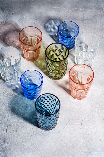 Colorful faceted and geometric drink glass, group of empty green, red, blue and transparent drinkware for beverage on stone concrete background, angle view