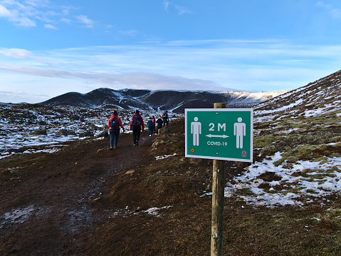 Grindavík, Iceland - 03-29-2021: People hiking on trail to recently erupted volcano at Fagradalsfjall mountain with warning sign reminding to keep two meters distance due to the Covid-19 pandemic.