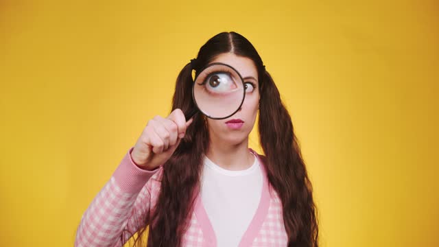 Young funny attractive caucasian girl with long braids holding a magnifying glass in front of camera and makes faces and grimaces on yellow backgorund. Human curiosity search concept
