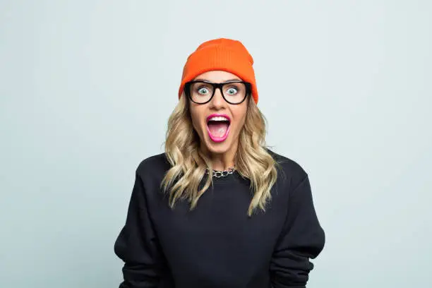 Excited young woman wearing street style black blouse and orange beanie hat, looking at camera and shouting.