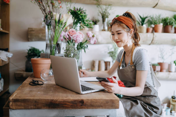 fioraio asiatico, proprietario di un negozio di fiori per piccole imprese, che utilizza lo smartphone mentre lavora su laptop contro fiori e piante. controllare le scorte, prendere ordini dei clienti, vendere prodotti online. routine quotidiana di gestione - florist telephone flower business foto e immagini stock