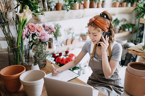 Asian female florist, owner of small business flower shop, talking on phone while working on laptop against flowers and plants. Checking stocks, taking customers orders, selling products online. Daily routine of running a small business with technology