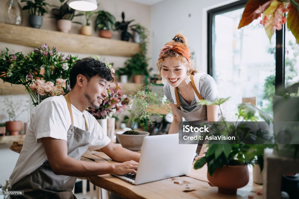 Smiling young Asian couple, the owners of small business flower shop, discussing over laptop on counter against flowers and plants. Start-up business, business partnership and teamwork. Working together for successful business Small Business Stock Photo