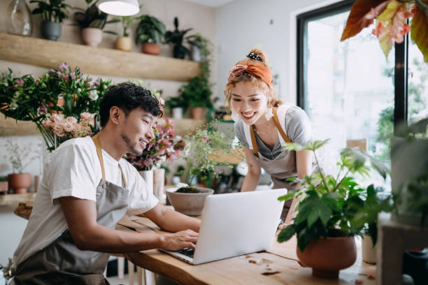 sorrindo jovem casal asiático, os donos de pequenas lojas de flores, discutindo sobre laptop no balcão contra flores e plantas. negócios iniciantes, parcerias comerciais e trabalho em equipe. trabalhando juntos para negócios de sucesso - best price fotos - fotografias e filmes do acervo